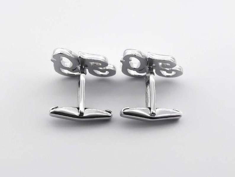 May include: A pair of silver cufflinks with the letters "DE" in a stylized font.