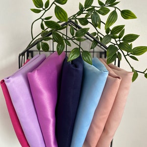 May include: A set of seven fabric squares in various colours, including pink, purple, blue, and peach, displayed in a black wire rack. The fabric squares are folded and arranged in a cascading pattern.