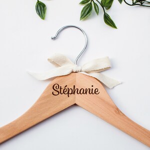 Peut inclure: Un cintre en bois avec le nom "Stéphanie" gravé en écriture cursive. Le cintre a une finition en bois naturel et est orné d'un nœud de couleur crème. Un crochet en métal argenté complète le design.