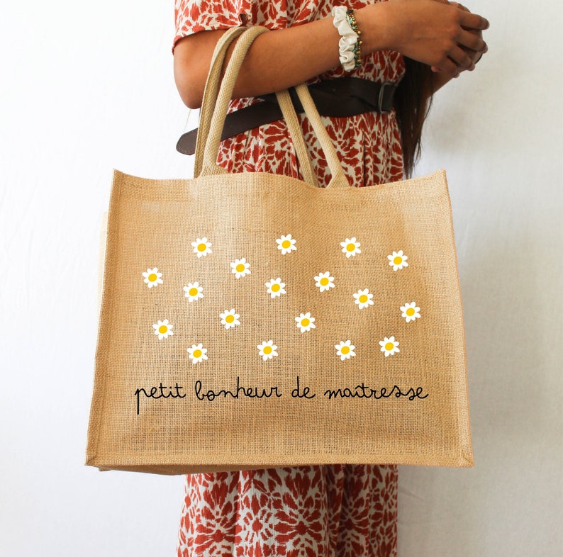 Pode incluir: Uma bolsa de juta marrom com margaridas brancas e o texto "petit bonheur de maitresse" em preto.