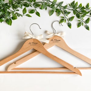 Peut inclure: Deux cintres en bois avec un ruban blanc et le texte "La Mariée" et la date "3/12/2022" gravés dessus.