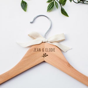 Peut inclure: Un cintre en bois avec un nœud en ruban blanc et le texte "JEAN & ELODIE" gravé sur le devant. Un petit motif de feuille se trouve sous le texte.