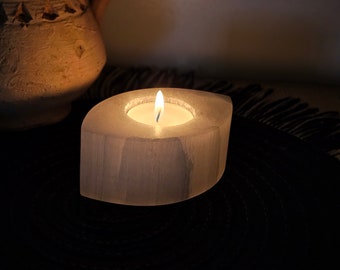 Può includere: Una candela accesa in un portacandele di selenite bianca a forma ovale. La fiamma della candela brilla intensamente sullo sfondo scuro. Un vaso decorativo color beige è sullo sfondo. Il portacandele è su una superficie nera strutturata.