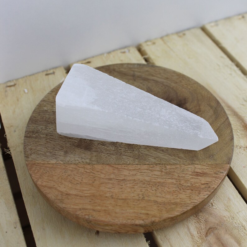 May include: A white selenite crystal wand, resting on a round, wooden coaster. The crystal has a triangular shape with a pointed end. The coaster is made of light and dark brown wood.