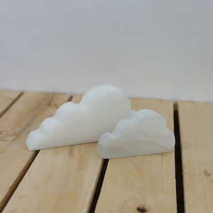 Puede incluir: Dos objetos decorativos blancos con forma de nube. Uno es más grande y redondeado, el otro más pequeño y plano. Se colocan sobre una superficie de madera marrón claro, con un fondo blanco liso. Las nubes parecen estar hechas de un material liso y sólido.