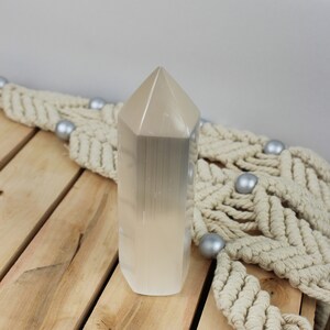 Puede incluir: Una alta torre de cristal de selenita de color claro con una parte superior facetada. El cristal se exhibe sobre una superficie de madera con un acento de macramé color crema y esferas decorativas plateadas.