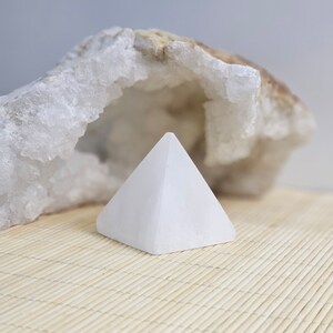 Könnte beinhalten: Eine kleine, weiße Pyramide aus Stein liegt auf einer hellen Bambusmatte. Dahinter befindet sich eine Geode mit rauer, strukturierter Oberfläche. Die Pyramide hat ein glattes, sauberes Aussehen.