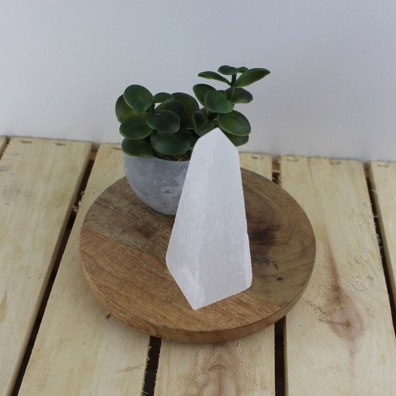 May include: A white selenite crystal tower stands on a wooden surface. The crystal has a smooth, polished appearance and a geometric shape. A small potted succulent plant sits behind the crystal. The background is a light wood surface.