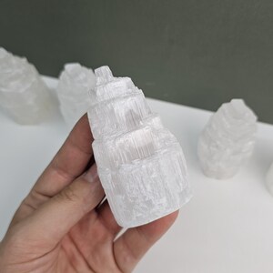 Può includere: Una mano che tiene una torre di selenite bianca, una formazione cristallina con un aspetto stratificato e strutturato. Altri pezzi di selenite sono visibili sullo sfondo. La torre è alta circa 10 cm.