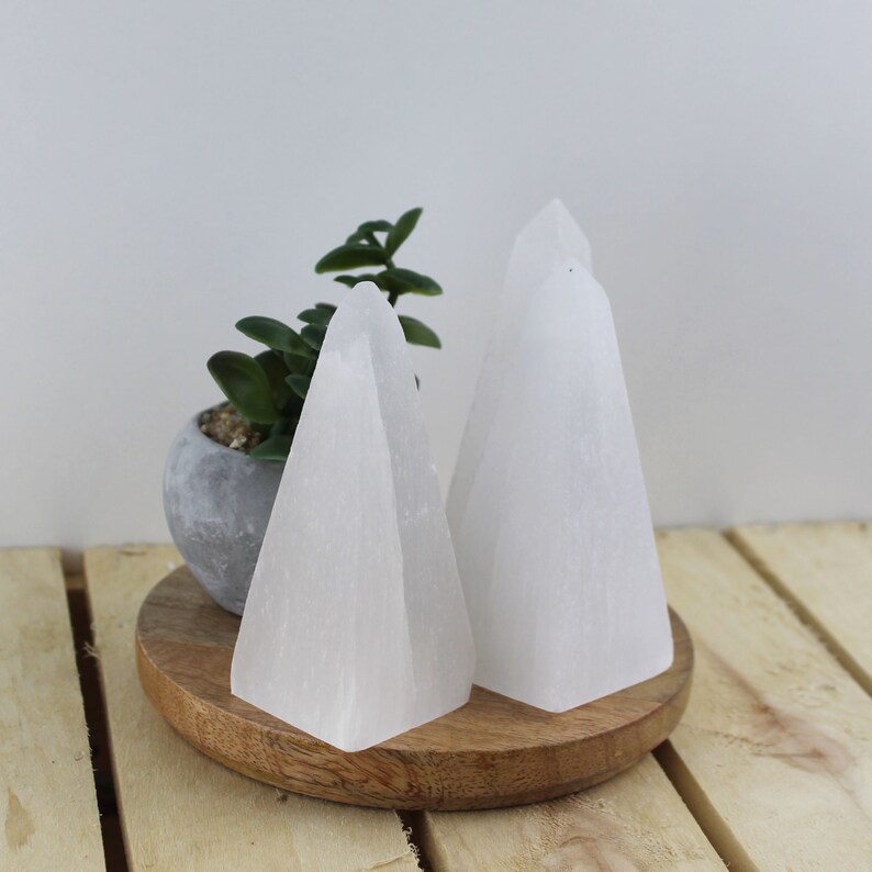 May include: Two white selenite pyramids of varying sizes are displayed on a wooden tray. A small potted plant with green leaves sits to the left of the pyramids. The pyramids have a smooth, polished appearance and are set against a light background.