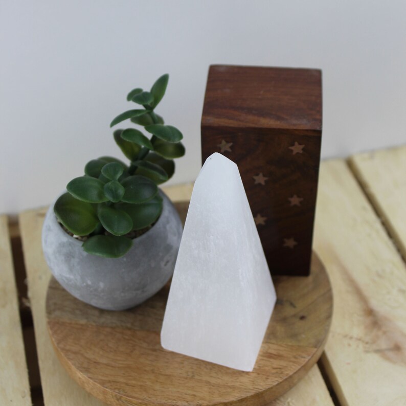 May include: A white selenite crystal pyramid stands on a wooden tray, next to a small potted succulent and a dark wooden box with star cutouts. The pyramid is the focal point, with a smooth, clean surface.