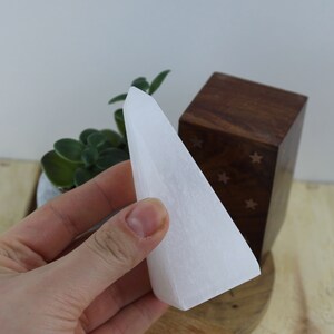May include: A white selenite crystal tower with a smooth, tapered shape. The crystal is held in a hand, with a wooden box and a small potted plant in the background. The tower is approximately 4 inches tall.