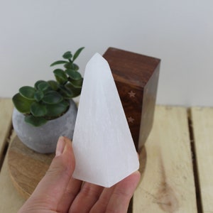 May include: A white selenite crystal tower with a triangular shape. The crystal is held in a hand, with a small potted plant and a wooden box in the background. The crystal is approximately 6 inches tall.