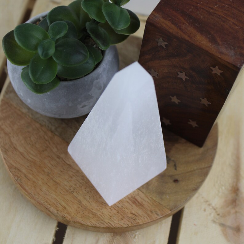 May include: A white, geometric crystal stands on a wooden tray, next to a small potted succulent and a wooden box with star cutouts. The crystal has a smooth, opaque surface and a pointed top.
