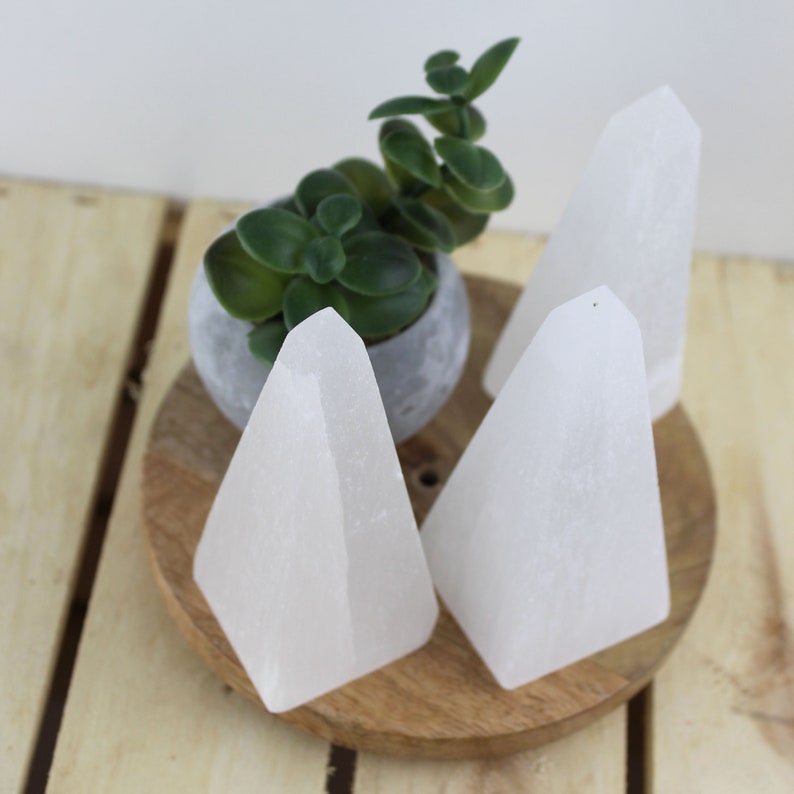 May include: Three white selenite crystal obelisks of varying sizes are displayed on a wooden tray. A small potted succulent plant in a gray pot sits behind the crystals. The background is a light wooden surface.