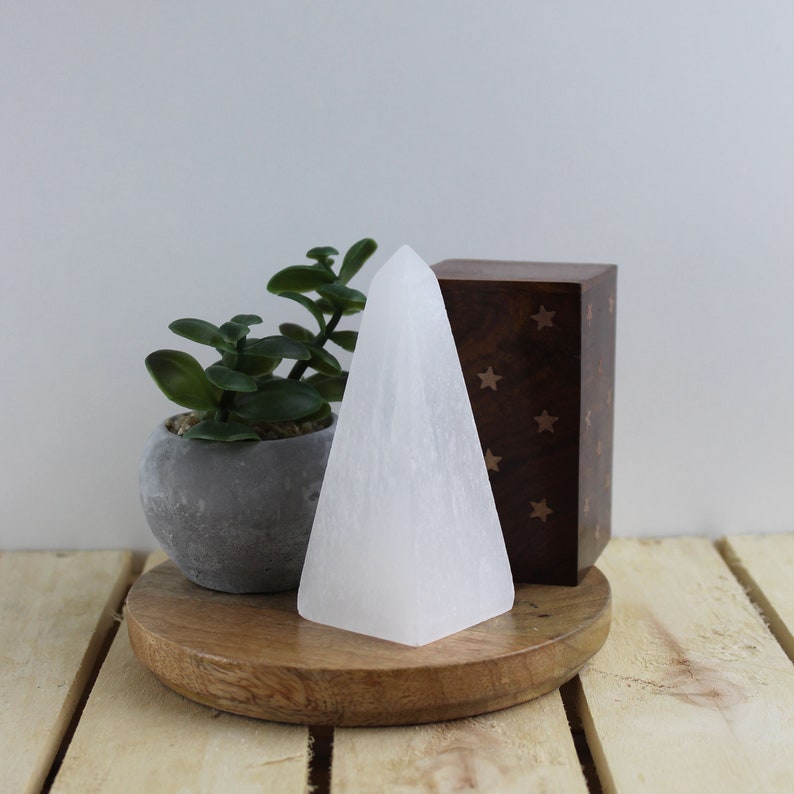 May include: A white, pyramid-shaped selenite crystal stands on a wooden surface. A small potted succulent and a wooden box with star cutouts are also present. The crystal is approximately 4 inches tall.