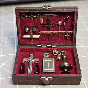 May include: Miniature wooden box with a red velvet interior, containing small religious items. Includes crosses, vials, a book with the text "Rituale Romanum", and other detailed accessories. The box is brown with a brass clasp.