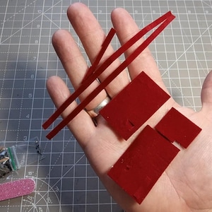 May include: A hand holding several small red rectangular pieces and two thin red strips. A small bag with miniature objects and a pink nail file are also visible.