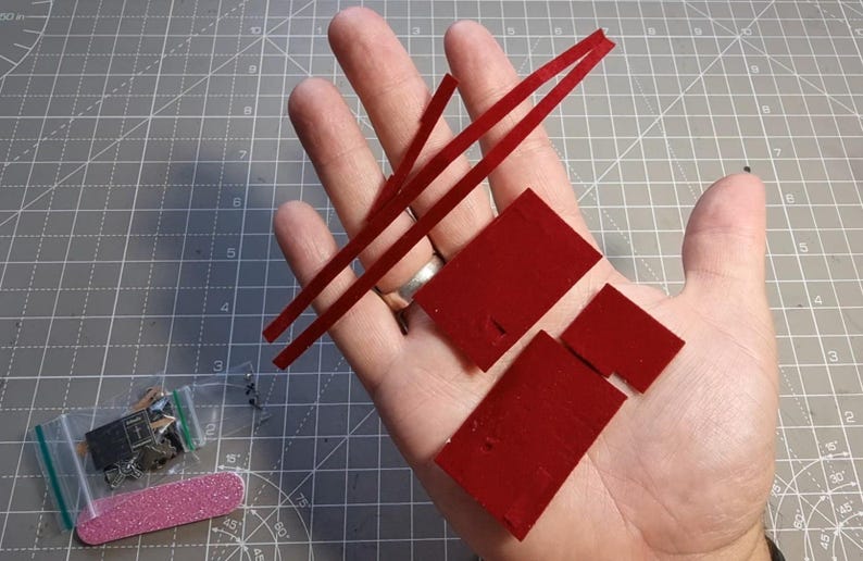 May include: A hand holding several small red rectangular pieces and two thin red strips. A small bag with miniature objects and a pink nail file are also visible.
