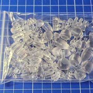 May include: A collection of miniature clear resin bottles, flasks, and goblets contained within a transparent plastic zip-top bag. The assortment includes various shapes and sizes of small containers, all featuring a smooth, translucent finish.