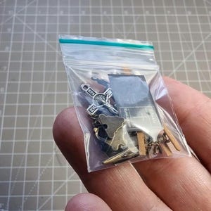 May include: A clear plastic bag containing various small metal charms. The charms include a cross, a sword, a map, and other shapes. The bag is sealed with a blue and green strip.