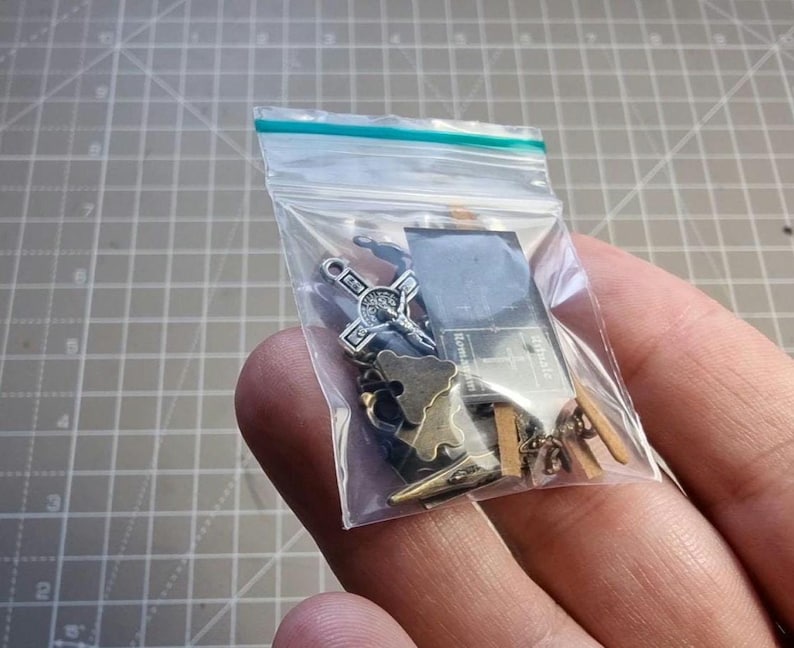 May include: A clear plastic bag containing various small metal charms. The charms include a cross, a sword, a map, and other shapes. The bag is sealed with a blue and green strip.