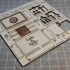 May include: A laser-cut wooden kit labeled "1:12th Scale Exorcism Kit." The kit includes various cut-out shapes, including a pentagram, cross, and other geometric forms. The text "Crafts By Xander UK 2025" is also visible.