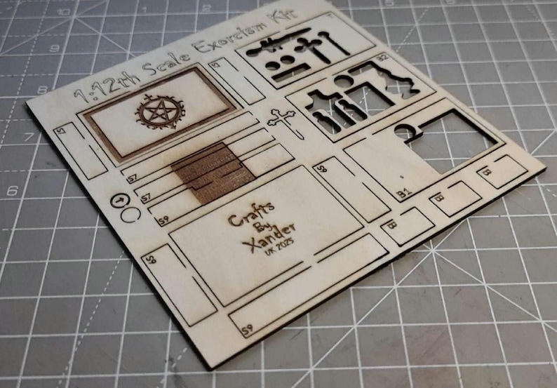 May include: A laser-cut wooden kit labeled "1:12th Scale Exorcism Kit." The kit includes various cut-out shapes, including a pentagram, cross, and other geometric forms. The text "Crafts By Xander UK 2025" is also visible.