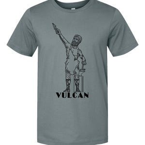 May include: A gray t-shirt featuring a black line drawing of Vulcan, the Roman god of fire and metalworking. The illustration depicts Vulcan holding a spear and hammer, standing next to an anvil. The word "VULCAN" is printed below the figure.