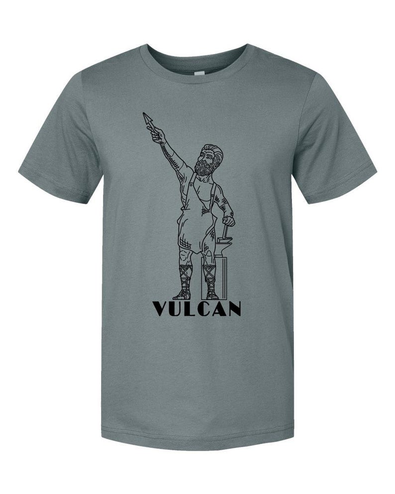 May include: A gray t-shirt featuring a black line drawing of Vulcan, the Roman god of fire and metalworking. The illustration depicts Vulcan holding a spear and hammer, standing next to an anvil. The word "VULCAN" is printed below the figure.