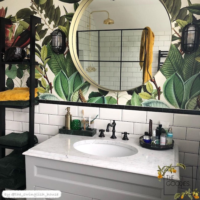 May include: A white bathroom vanity with a marble countertop and a white porcelain sink. The vanity has a black framed mirror with a gold trim and a tropical leaf wallpaper behind it. The vanity has a black metal shelf with towels on it. The vanity has a black metal faucet and a black metal soap dispenser.