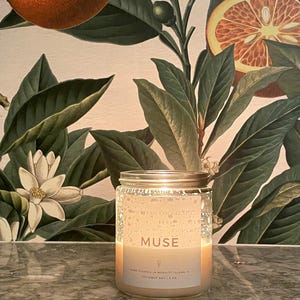 May include: A lit glass jar candle with a silver lid. The candle has a white label with the word "MUSE" and other text. The background features a botanical print with oranges and green leaves. The candle is 8 oz.