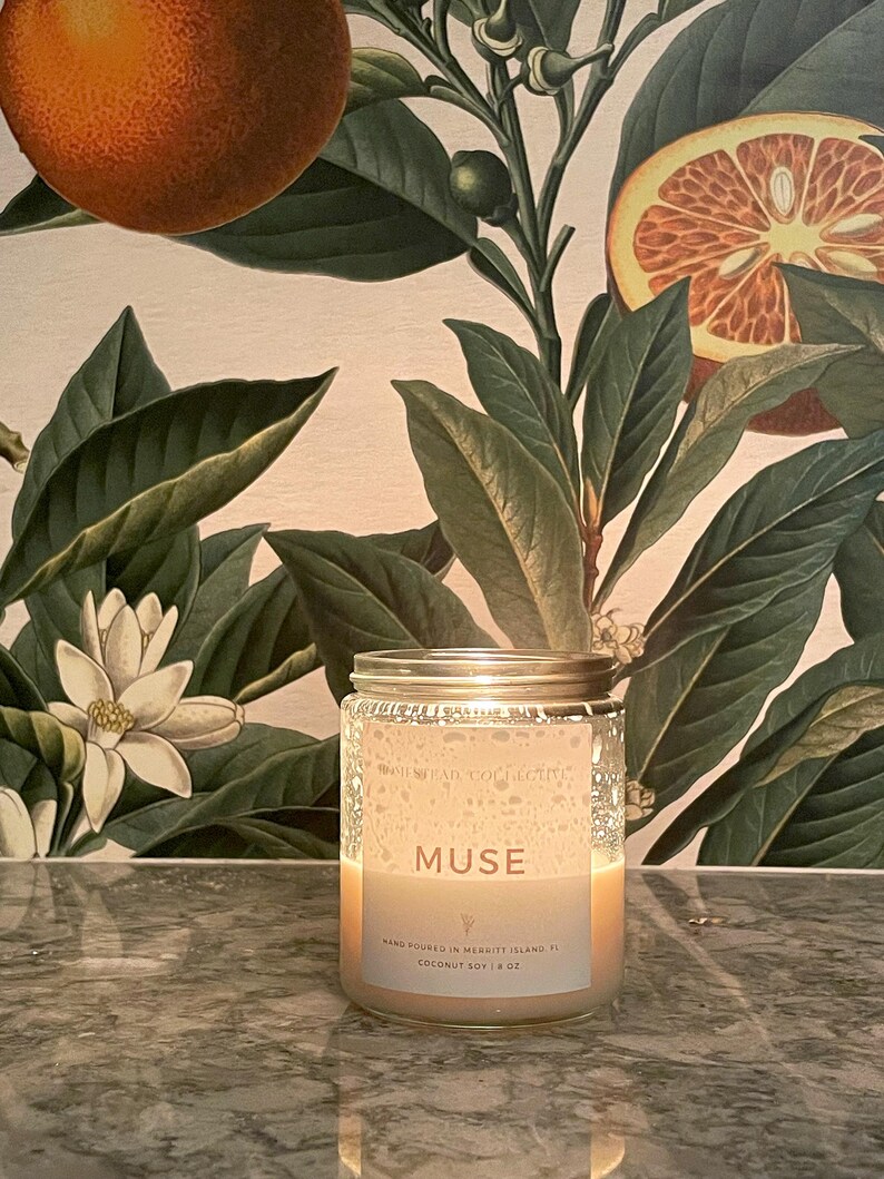 May include: A lit glass jar candle with a silver lid. The candle has a white label with the word "MUSE" and other text. The background features a botanical print with oranges and green leaves. The candle is 8 oz.