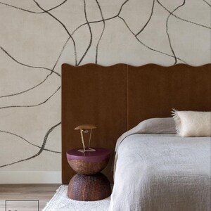 Op de afbeelding: Slaapkamerinterieur met een bruin hoofdbord met een golvende rand en een bed bedekt met een grijze deken. Een nachtkastje met een donkerpaarse bovenkant en geweven basis staat op een wit tapijt. De muur is beige met zwarte lijnen.