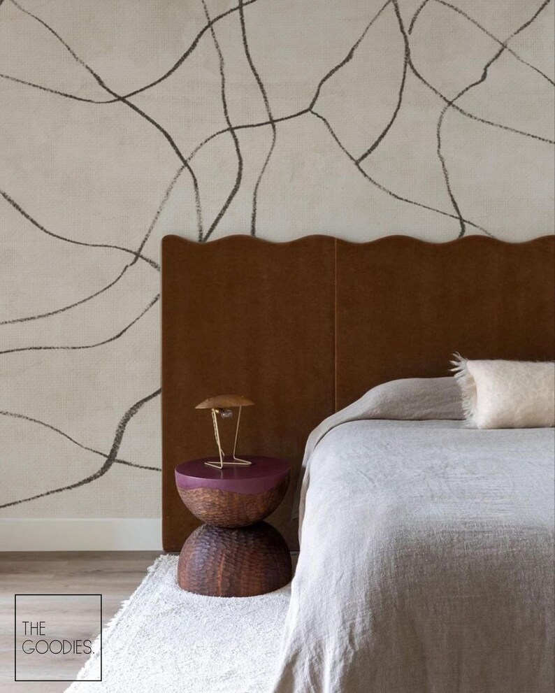 Op de afbeelding: Slaapkamerinterieur met een bruin hoofdbord met een golvende rand en een bed bedekt met een grijze deken. Een nachtkastje met een donkerpaarse bovenkant en geweven basis staat op een wit tapijt. De muur is beige met zwarte lijnen.