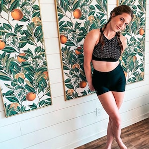May include: A woman wearing a black sports bra with a geometric pattern and black shorts. Behind her are three framed botanical prints featuring oranges, green leaves, and white flowers against a cream background.