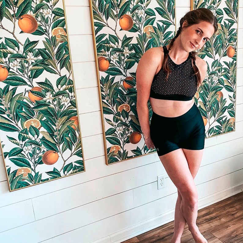 May include: A woman wearing a black sports bra with a geometric pattern and black shorts. Behind her are three framed botanical prints featuring oranges, green leaves, and white flowers against a cream background.