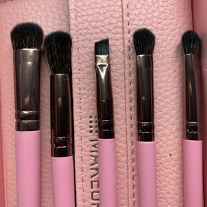 Puede incluir: Un juego de cinco brochas de maquillaje con cerdas negras y mangos rosas. Las brochas están dispuestas en un estuche de cuero rosa. Los mangos son de color rosa claro y las férulas metálicas son de color plata oscura.