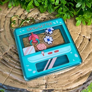 May include: A teal blue acrylic keychain shaped like a claw machine with the text "Grow Getter" on the front. The claw machine has a pink flower, a white flower, and a white flower with a blue center inside.