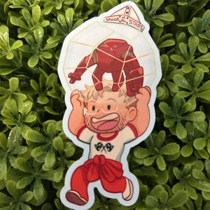 May include: A die-cut sticker featuring a cartoon character with blonde hair and glasses wearing a white t-shirt with a checkered flag graphic and red pants, holding a red robot inside a geometric cage with the text Savior of the Stars.