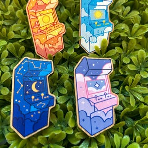 May include: Four colorful arcade machine pins. Each pin features a different sky-themed design: sunset, daytime clouds with sun, starry night with moon, and a pink and purple sky. The pins are set against a backdrop of green foliage.