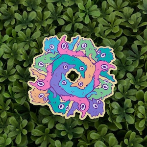 May include: A colorful sticker featuring a circular design of cartoonish creatures in shades of pink, purple, blue, green, and orange. The sticker has a yellow border and is set against a backdrop of green foliage.