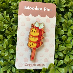 May include: A wooden pin featuring a stack of three cartoon bees with red and yellow accents. The pin is on a white card with the text "Wooden Pin" and "Cozy Gremlin".