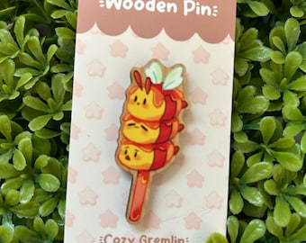 May include: A wooden pin featuring a stack of three cartoon bees with red and yellow accents. The pin is on a white card with the text "Wooden Pin" and "Cozy Gremlin".