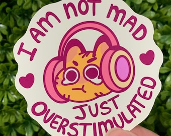 May include: A sticker with a cartoon cat wearing pink headphones and crying. The text on the sticker reads "I am not mad just overstimulated".