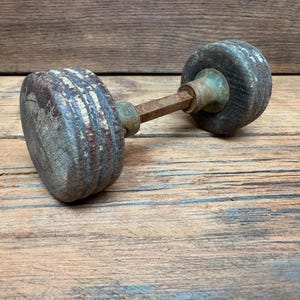 May include: A vintage door knob set featuring two circular, weathered wooden knobs with visible grain and texture, connected by a central square metal spindle. The hardware includes aged brass-toned circular collars at the base of each wooden knob.