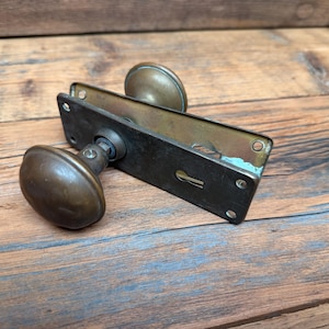 May include: A vintage brass door handle set featuring two rounded knobs attached to a rectangular backplate. The metal has a tarnished, aged patina with visible screw holes at the corners and a central keyhole slot on the rectangular plate.