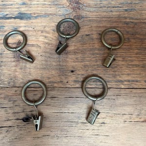 Op de afbeelding: Vijf ronde gordijnringen van antiek messingkleurig metaal, elk voorzien van een bevestigde metalen clip met veer. De ringen zijn verspreid gerangschikt, waardoor de metalen afwerking en het functionele ontwerp van de clips goed zichtbaar zijn.