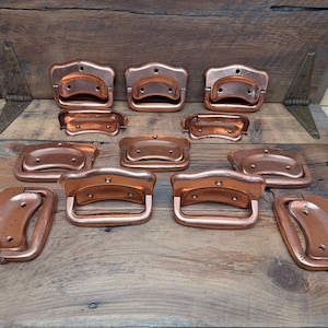 May include: A collection of ten copper-colored metal drop handles arranged in rows. Each handle features a rectangular backplate with rounded corners and two screw holes, paired with a curved, hinged pull mechanism for furniture attachment.