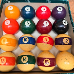 May include: A set of fifteen numbered billiard balls arranged in a box. The top row features solid yellow, blue, red, and purple balls. The second row includes solid orange, green, maroon, and black balls. The bottom rows contain striped balls.